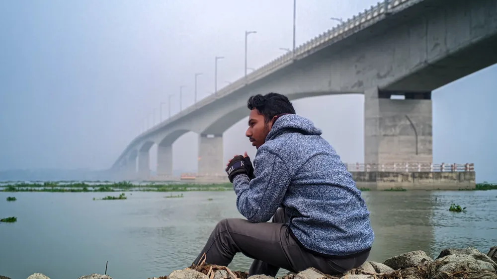 Sefat Khan sitting on rocks by the water with a long bridge in the background